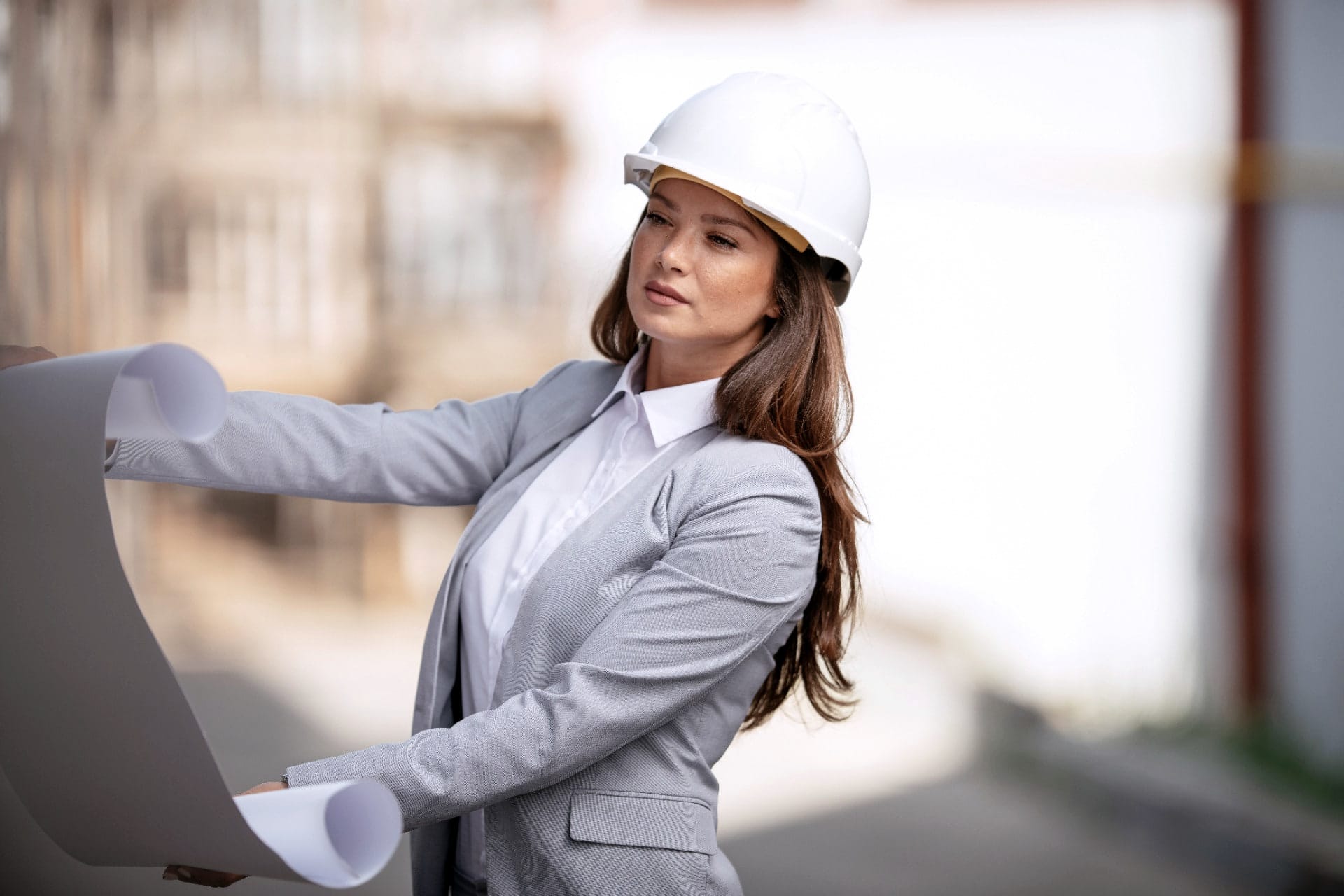 A professional in a white hard hat and grey jacket studies a large drawing on site.